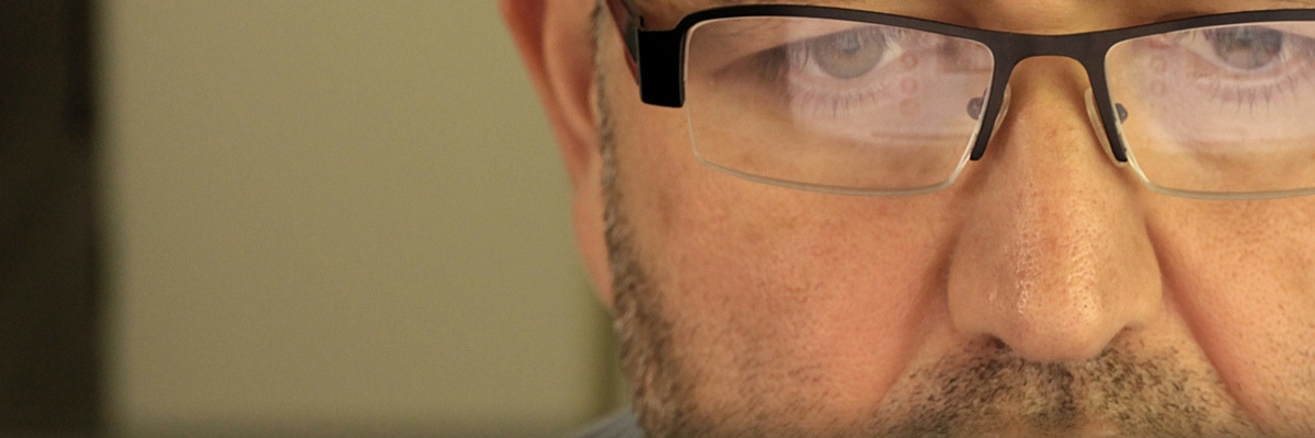 Un homme vue de face devant un ordinateur visible par le reflet de l'écran dans ses lunettes